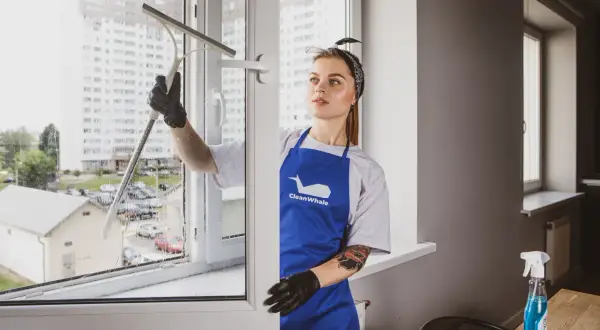 Kobieta w niebieskim fartuchu myje okno przy użyciu ściągaczki.
